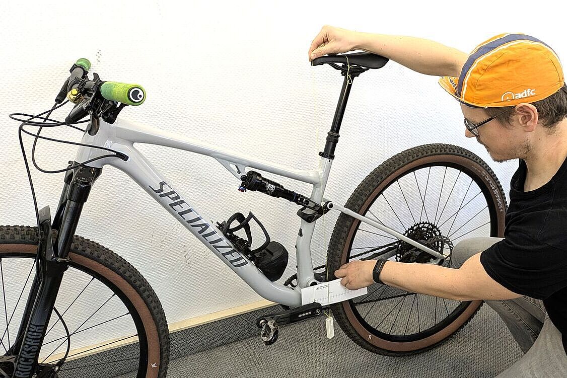 Ein Mann kniet vor einem Mountainbike und hält ein Senkblei an die Sattelnase und eine Schablone an die Kurbel. 