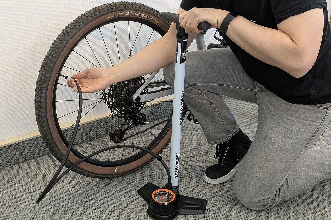 Ein Mann kniet vor einem MTB-Hinterrad und steckt den Pumpenkopf einer Standpumpe auf das Ventil. 