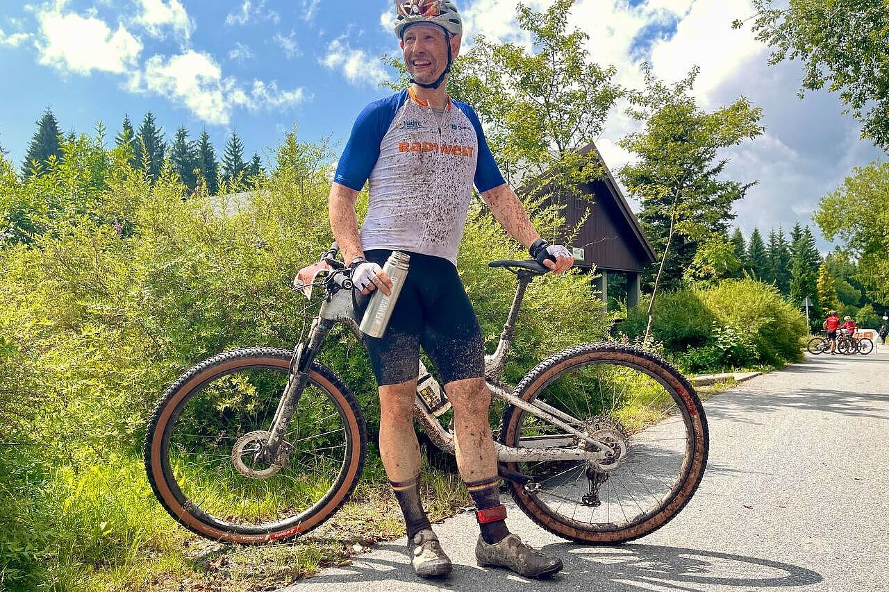 Im Ziel Ein mit Schlamm bespritzter Mountainbiker sitzt auf dem Oberrohr seines MTB und hält eine Trinkflasche in der Hand.