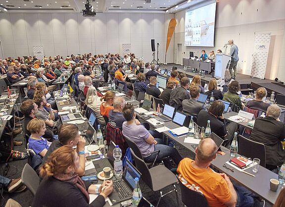Ein grpßer Saal mit 140 Menschen an Tischreihen, die zur Bühne blicken, wo Boris von Heesen einen Vortrag hält. 