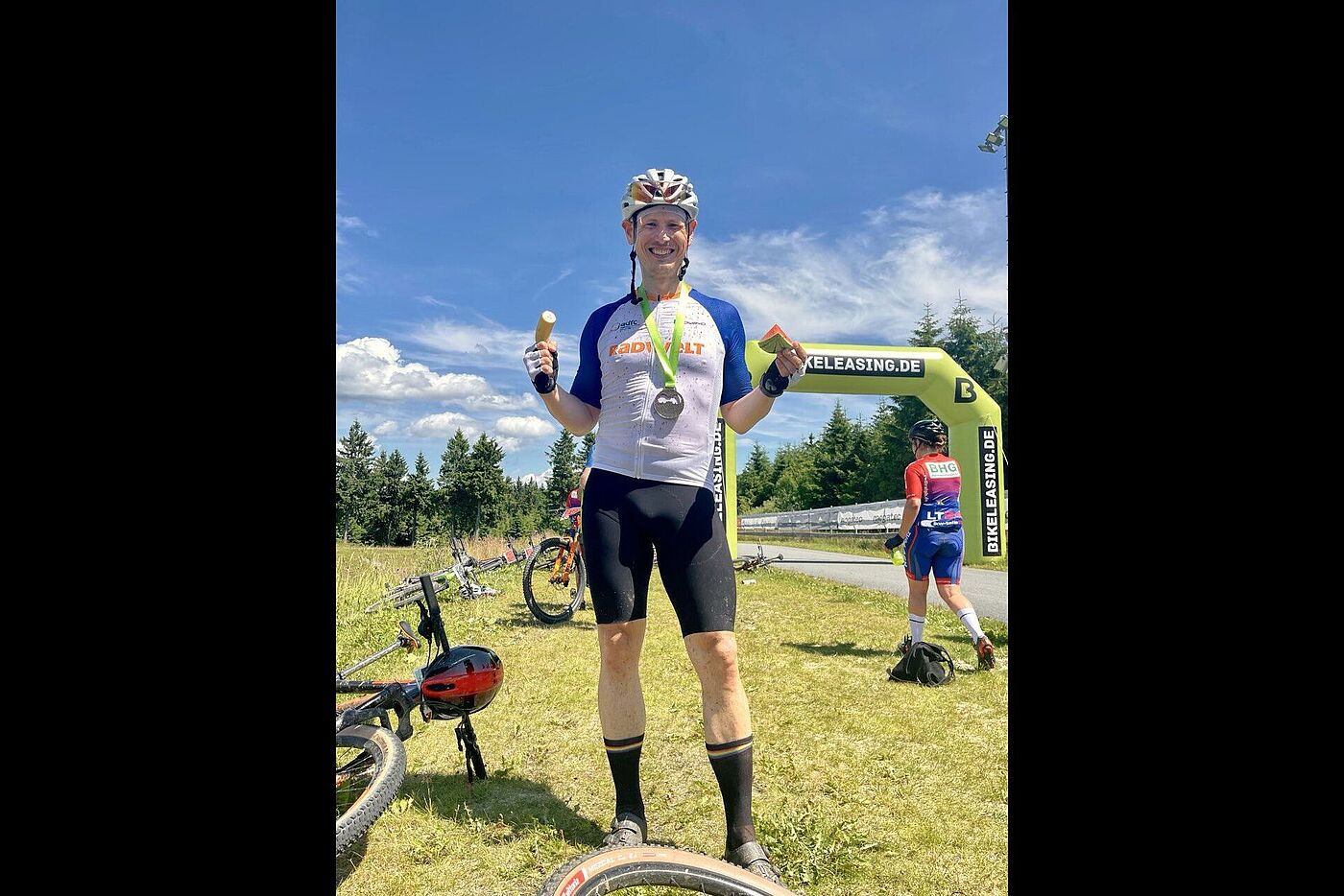 Medaille Ein Mountainbiker steht lachend im Zielbereich eines MTB-Rennens, um den Hals trägt er eine Finisher-Medaille, in den Händen hält er eine Banane und ein Stück Melone.