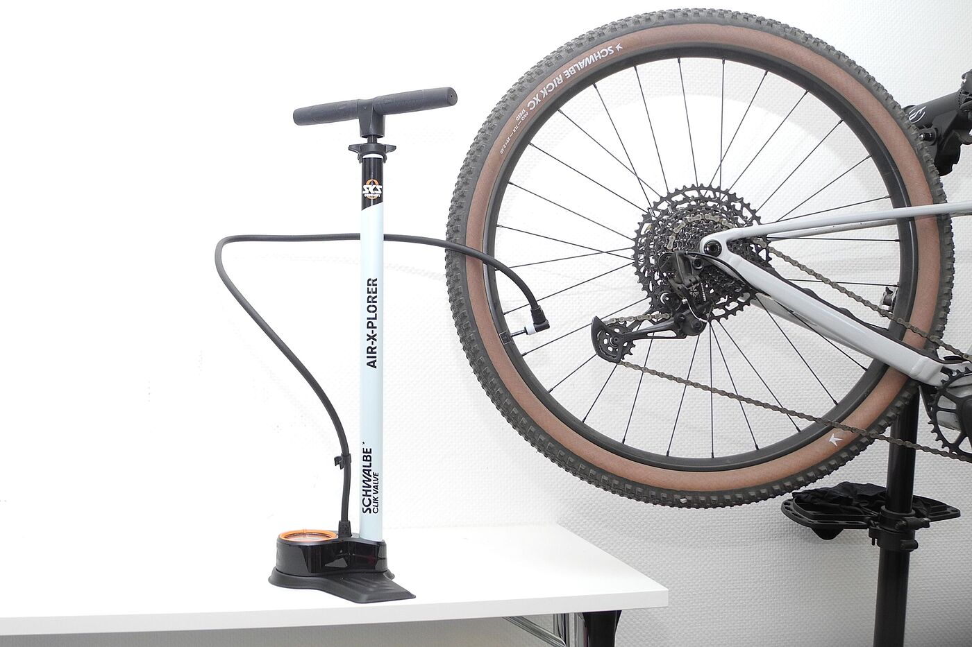 Eine Standpumpe steht auf einem Tisch vor weißem Hintergrund, der Schlauch führt zu einem MTB-Hinterrad. 