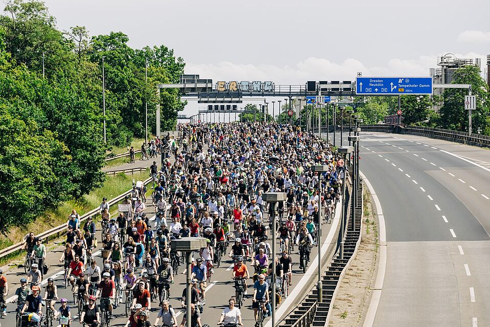 Sternfahrt Berlin 2025 Eine große Menschenmenge fährt auf Fahrrädern über eine mehrspurige Autobahn, die bis zum Horizont mit Radfahrenden gefüllt ist. Über der Straße befinden sich Verkehrsschilder, die unter anderem Richtung „Dresden“ und „Neukölln“ zeigen. Links säumen Bäume die Straße, rechts ist eine leere Gegenfahrbahn zu sehen.